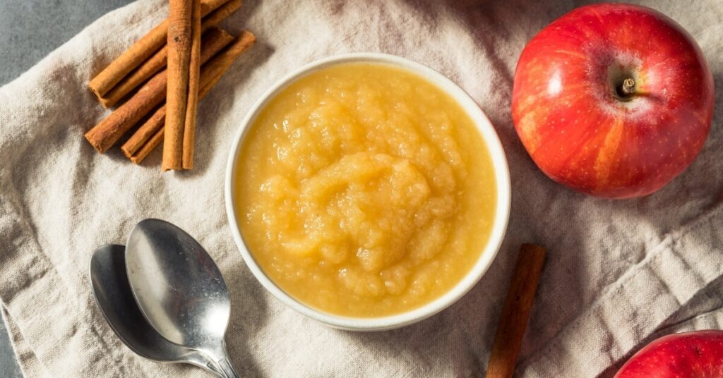 image representing a bowl full of applesauce alongside an apple and cinnamon